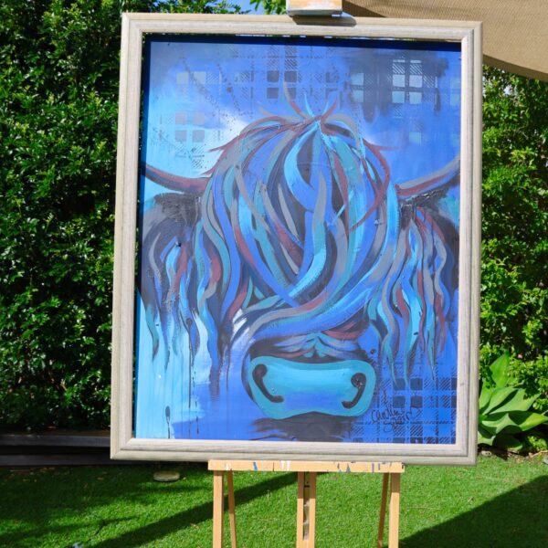 Ally, an original blue Highland cow painting by Caroline Speirs in a pale silver frame, on an easel in her Perth garden