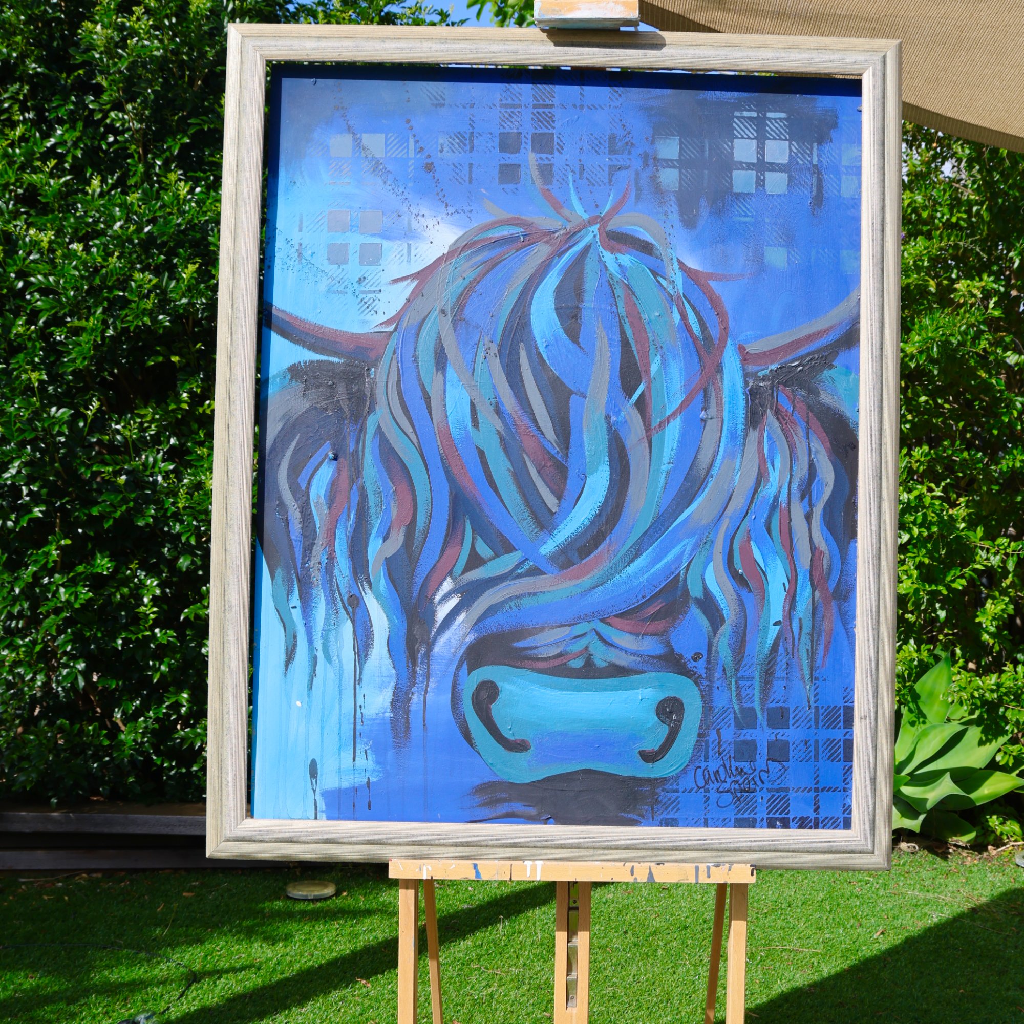 Ally, an original blue Highland cow painting by Caroline Speirs in a pale silver frame, on an easel in her Perth garden
