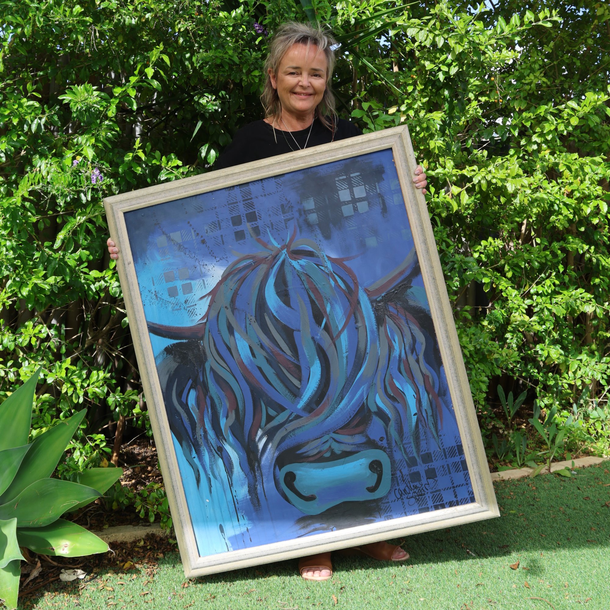 Caroline Speirs holding Ally, an original blue Highland cow painting in a pale champagne silver frame, in her Perth garden