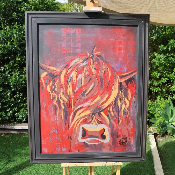 Fiona, an original red Highland cow painting by Caroline Speirs in a black frame, on an easel in her Perth garden