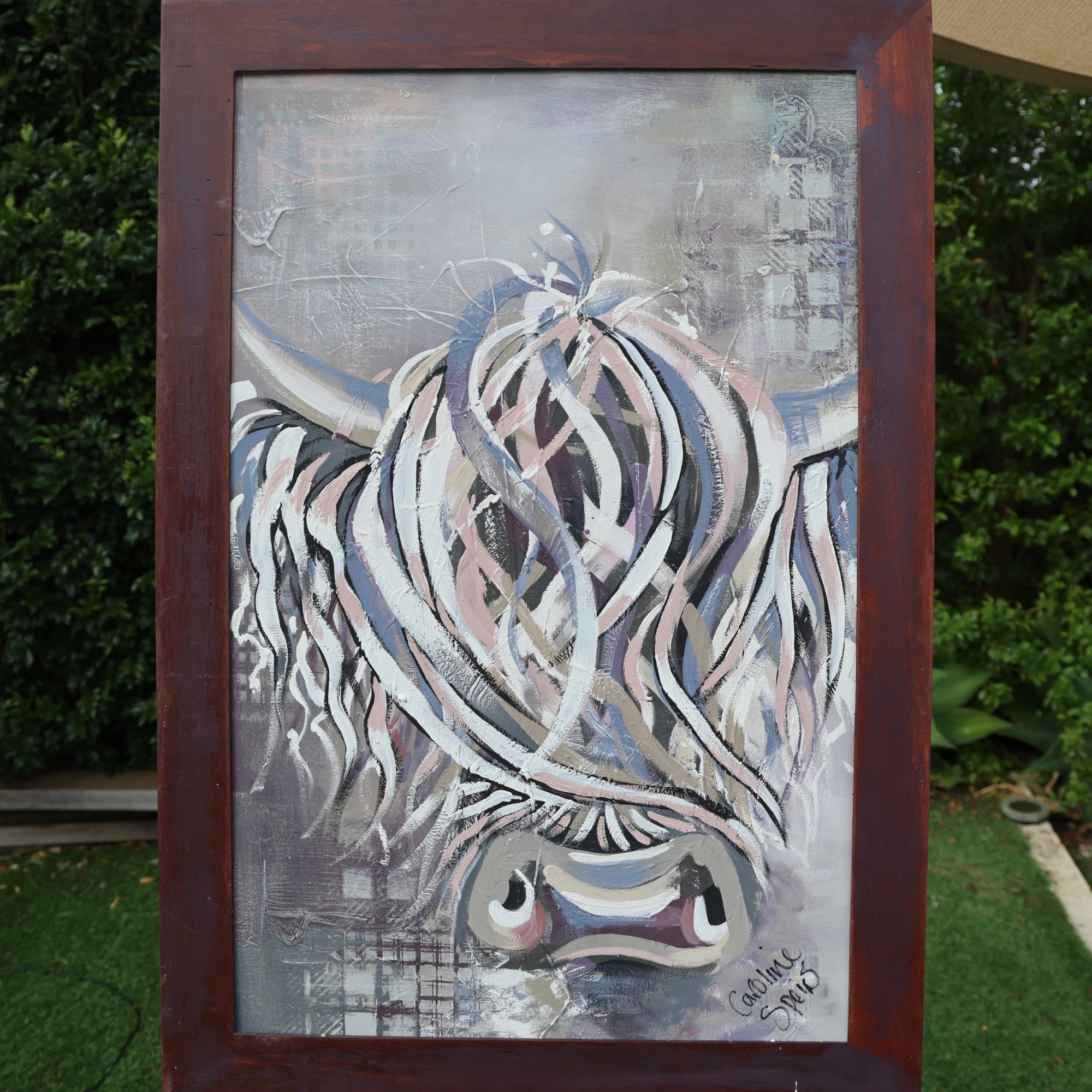 Isla, an original Highland cow painting in white, dusky pink, slate blue and mauve by Caroline Speirs, in a dark red timber frame on an easel in her Perth garden