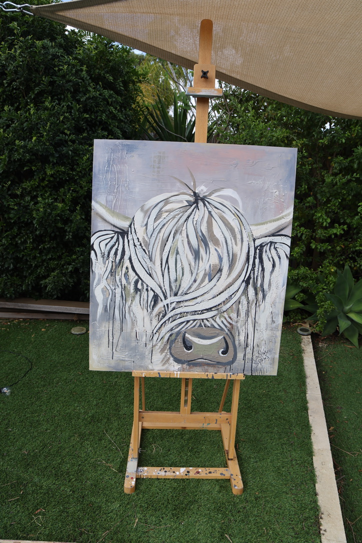 Moira, an original white and blush pink Highland cow painting by Caroline Speirs, unframed on a wooden easel in her Perth garden