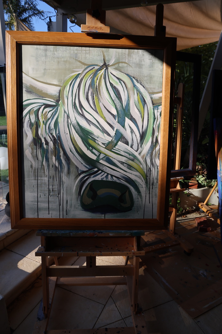 Paddy, an original green Highland cow painting by Caroline Speirs, framed in oak on an easel in the late afternoon sun